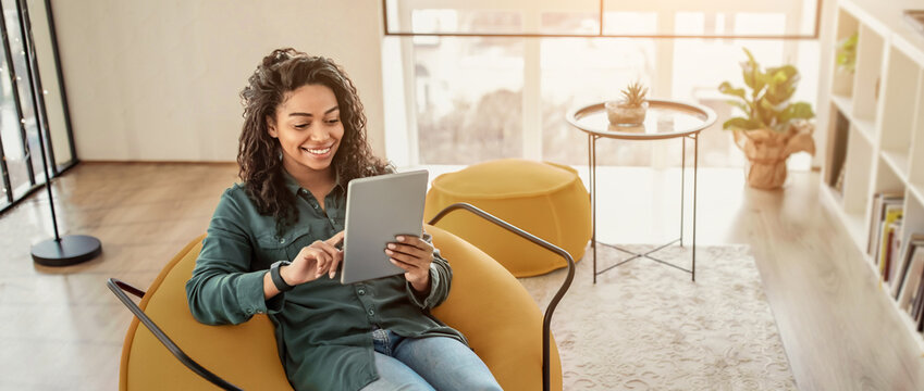 A young woman smiles while sitting comfortably in a modern yellow chair. She is using a tablet in a bright living area with plants and a coffee table nearby.