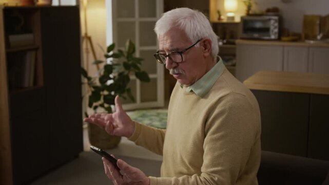 Medium arc shot of senior 80-year-old Caucasian man sitting on couch at home, revealing online bank app password to cybercrime fraudster, getting money transfer alert and shaking fists with rage