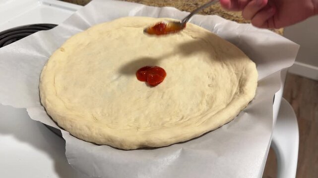 Fresh pizza base topped with tomato sauce on parchment paper, prepared at home and ready for cheese and baking.