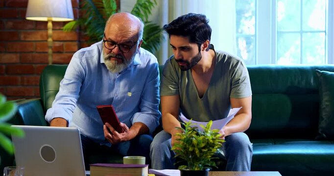 Indian retired father and young son celebrating online business success while working on laptop and smartphone at home, reviewing financial report and share trading profit with joyful family moment