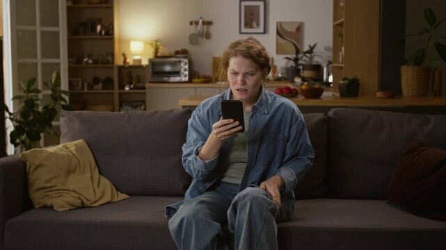 Full zoom shot of young Caucasian female cyber scam victim relaxing on couch at home, seeing money transfer alerts on smartphone, sobbing and yelling, after revealing password on phishing website