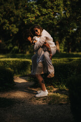 Fototapeta premium A couple shares a playful piggyback moment in a sunlit park. They smile and laugh as one carries the other along a garden path.