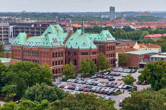 Helsingborg, Sweden - 12 August 2025: View of the copper-green roofs of a grand red-brick building amidst lush trees and a full parking lot, set against a backdrop of the city skyline.