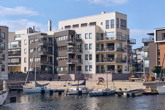 Landskrona, Sweden - 08 August 2025: View of modern buildings reflect against the serene harbor waters, where small boats bob gently beneath a bright, clear sky.