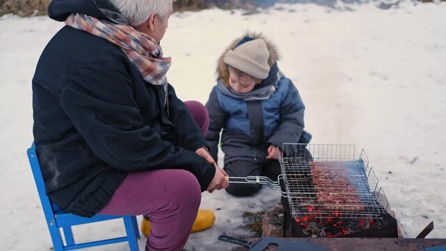 Gray haired grandfather and child by grill roasting sausages on charcoal brazier, bundled in winter coats and scarves on snowy ground. Hands warmed over glowing embers, smoke. Family outdoor barbecue.
