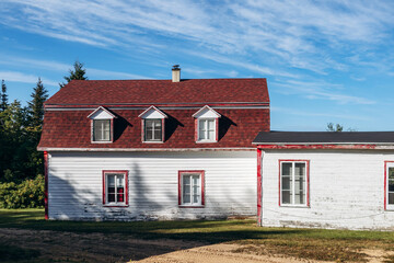 Obraz premium Historic white wooden house with red trim at Saint Firmin Historic Village, traditional Quebec rural architecture with church steeple visible behind the building under blue summer sky