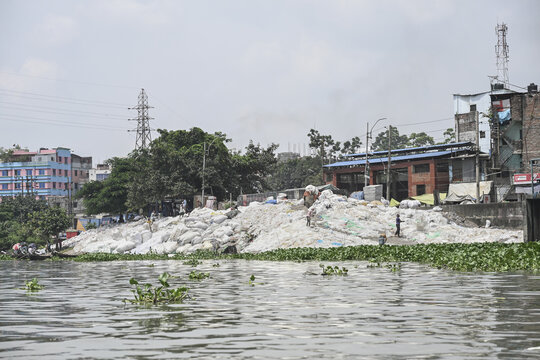Dhaka, Bangladesh - 10 September 2025: View of a riverbank piled high with white plastic waste against a backdrop of buildings and green vegetation.