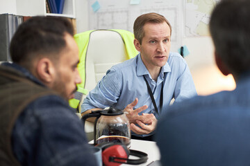 Middle aged Caucasian man sitting at desk discussing project details with two male colleagues in...