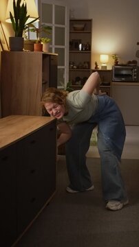 Full vertical shot of mature Caucasian woman moving and rearranging furniture in living room, grabbing cabinet, then collapsing with lower back injury, crying in pain and massaging lumbar area