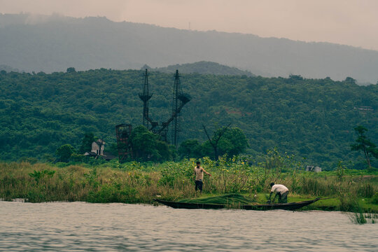Bholaganj, Bangladesh - 27 August 2021: View of verdant hills meeting the tranquil waters where workers harvest the river's bounty under a hazy sky.
