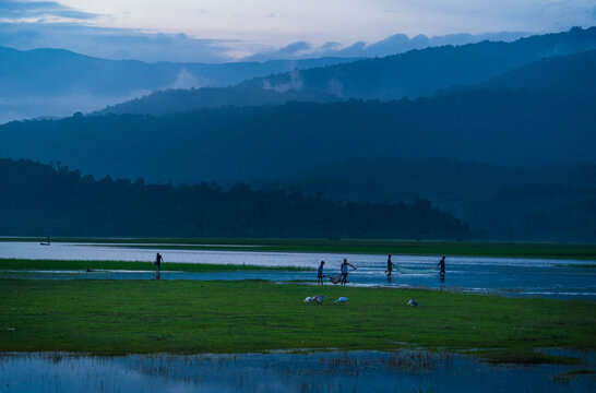 Bholaganj, Bangladesh - 27 August 2021: View of figures wading through the shallow waters amidst the lush green landscape, framed by the majestic, misty mountains in the distance.