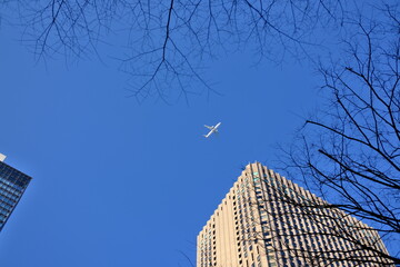 都会のビル群に囲まれた広大な青空を横切る白い機体
