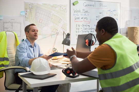 Caucasian middle aged man sitting at desk handing documents to Black middle aged man wearing safety vest, both discussing construction project in office with blueprints on wall
