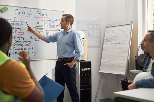 Caucasian middle aged man standing in office pointing at whiteboard with construction plans, while multiethnic group of building workers listening and taking notes during meeting