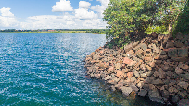 A serene rocky shoreline with lush greenery meets a vast body of water under a blue sky