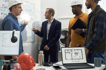 Plakat Group of middle aged men of diverse ethnicities discussing construction plans in office, one Caucasian man pointing at whiteboard while others listening, laptop displaying building floor plan