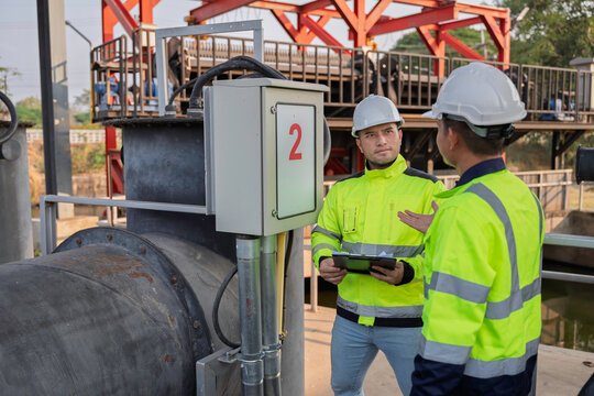 Two Asian engineers inspecting electrical control cabinet at irrigation waste trap station. Professional technicians checking water management system and monitoring hydraulic equipment outdoors.