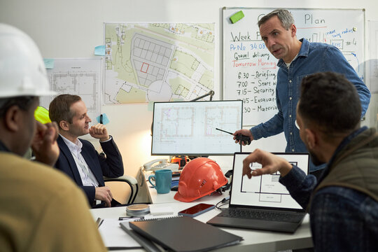 Middle aged Caucasian man standing and gesturing with pen while leading meeting with diverse group of middle aged men in office, discussing construction plans displayed on monitors