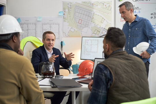 Caucasian middle aged man leading discussion with Black middle aged man, Middle Eastern young adult man, Caucasian middle aged man in office, construction plans and hard hats visible