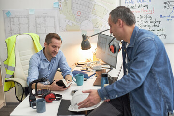 Middle aged Caucasian man sitting at desk reviewing documents with middle aged Caucasian man...
