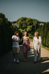Three friends walk along a sunny path, smiling and chatting. The scene captures a carefree, outdoor moment with a warm, sociable vibe.