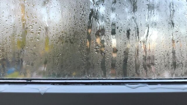 Water drops sliding down window glass, water trails forming on wet surface with condensation. Closeup of droplets pattern on transparent glass, blurred city view behind, natural background concept.