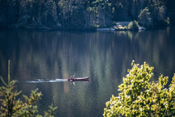 A lone person paddling a red canoe across the calm waters of the Saguenay River, surrounded by dense boreal forest and reflected trees