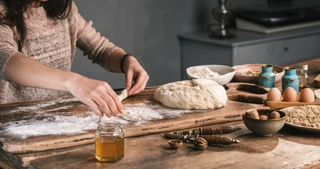 Fototapeten Brot In a cozy kitchen, a person carefully shapes dough on a wooden table, surrounded by flour, eggs, and jars of honey, creating a special bread from scratch  © Damian