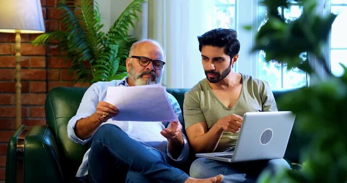 Indian retired father and young son working together on laptop and smartphone at home, reviewing financial report, share trading data and online business documents while discussing profit and loss
