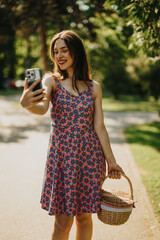 A smiling woman in a pink and blue floral dress takes a selfie in a sunny park. She carries a...
