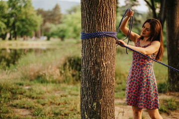 A woman in a floral dress ties a rope around a tree in a park. Outdoor scene shows greenery, sunlit grass, and calm, casual activity.