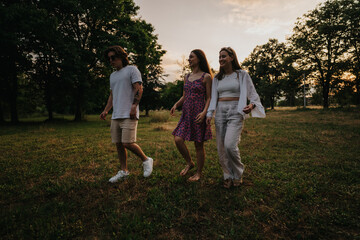 Fototapeta premium Three friends walk through a grassy park at sunset, smiling and relaxed. A warm outdoor scene suitable for lifestyle and friendship imagery.