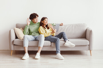 Obraz premium Cheerful millennial Asian couple using tablet for online communication, sitting on sofa, waving at webcam at home, free space. Young spouses having video chat with friend, enjoying remote conversation