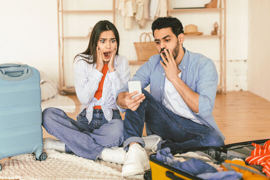 Oh no, flight is cancelled. Shocked spouses using cellphone and reading shocking travel news or negative message about tourism, sitting near suitcases at home