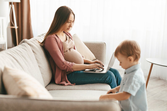 Pregnant young woman working on laptop computer while spending time with her little son at home, boy playing with toys near mom, woman sitting on sofa