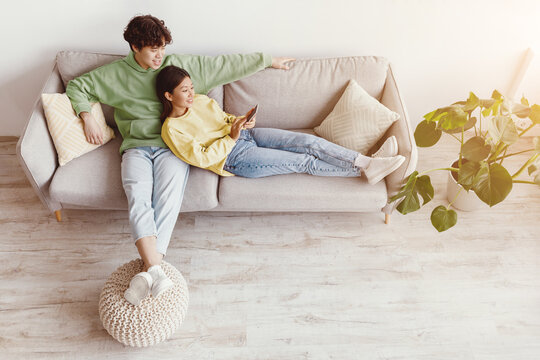 Loving Asian couple using smartphone, surfing internet or watching movie online, resting on sofa in living room, top view. Young husband and wife with mobile device chilling at home. Free space