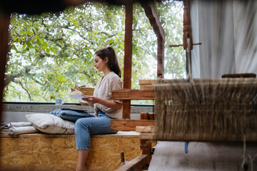 Fototapeta premium Young businesswoman making a call with customer, sitting by wooden loom. Upcycled textile, small weaving business and circular economy.