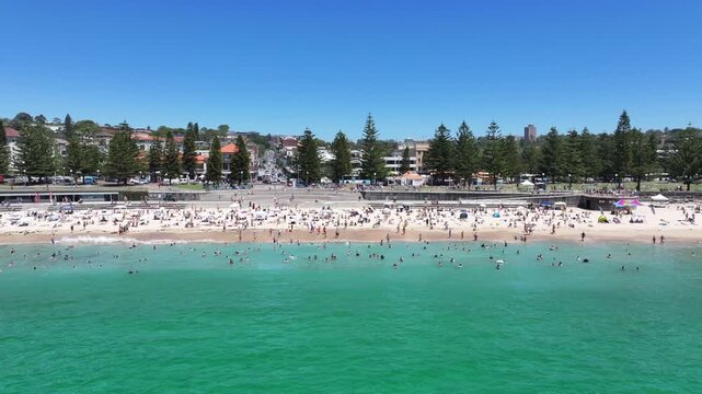 flying parallel and away from Coogee Beach, with many people sunbathing at the beach and Sydney cityscape in background, located in Coogee Bay, Pacific Ocean, Australia - 4k aerial video footage