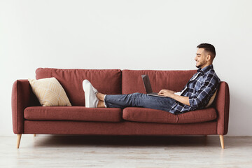 Fototapeta premium A man sits on a sofa with a laptop. He looks focused while typing. The living room is simple with a light background and a cushion on the couch. It is daytime.