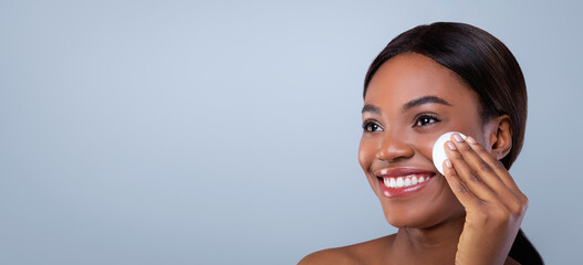 A woman is using a cotton pad to apply skincare product on her face. She has a bright smile and is standing indoors against a light blue wall. The room is simple and uncluttered. © Prostock-studio