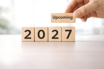 Naklejka premium Hand adding 'Upcoming' block to wooden blocks spelling '2027', representing future planning and New Year anticipation.