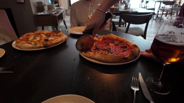 Unrecognizable person using a pizza cutter to slice a fresh neapolitan pizza with tomato sauce and anchovies while dining with a friend at a modern restaurant with glasses of craft beer