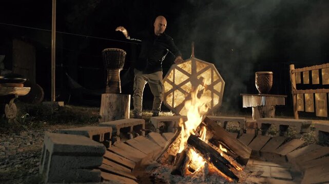 A spiritual native man playing a djemba drum by a burning fire at night. camera movement