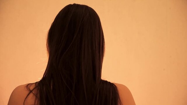 Young 30s sexy woman holds her long dark hair in a bun then lets it go while continuing to touch it. Rear view showing head, neck and shoulders. Morning rituals. Evening body bliss. Arousal and lust.