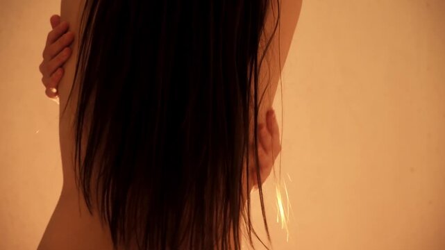 Rear view on back of young naked sexy woman with long dark hair sensuality touching her waist with her fingers. Studio lighting. Body bliss. Arousal and lust. Intimate and caring gesture. Foreplay.