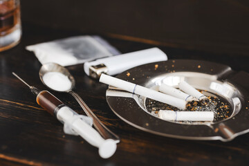 Concept of substance abuse and drug addiction, ashtray with cigarettes, lighter, plastic bag with powder, spoon and syringe with dose on dark wooden background