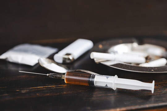 Concept of substance abuse and drug addiction, syringe with dose, ashtray with cigarettes, lighter, plastic bag with powder and spoon on dark wooden background