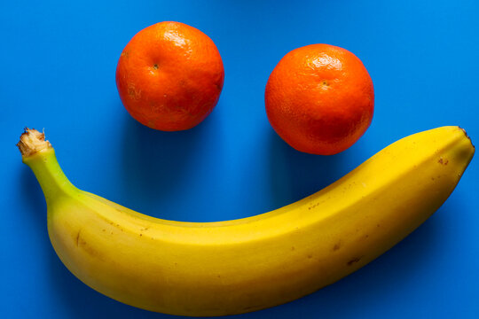 ​A composition of a bright yellow banana and two orange tangerines forms a positive smiling face on a rich blue background.
