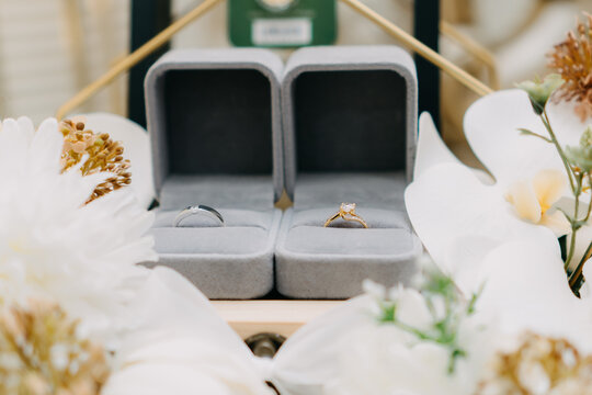 Pair of Wedding Rings in Velvet Boxes with White Flowers.Close-up of a silver men's band and a gold diamond engagement ring in grey velvet boxes surrounded by orchids.