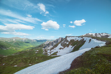 Scenic view from grassy flowering cliff along ridge with snow cornice to sharp rocks and rocky sheer crags under lush clouds in blue sky. Sunlight and shadows in high mountains in changeable weather. © Daniil